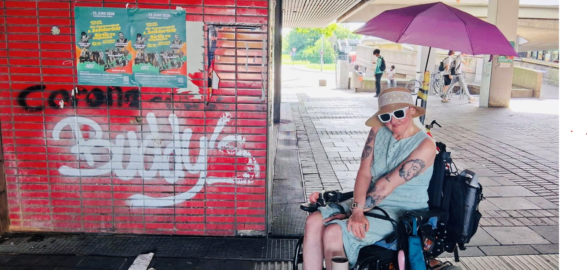 Eine Frau im Rollstuhl vor einer Wand mit Buddy-Graffiti in Köln